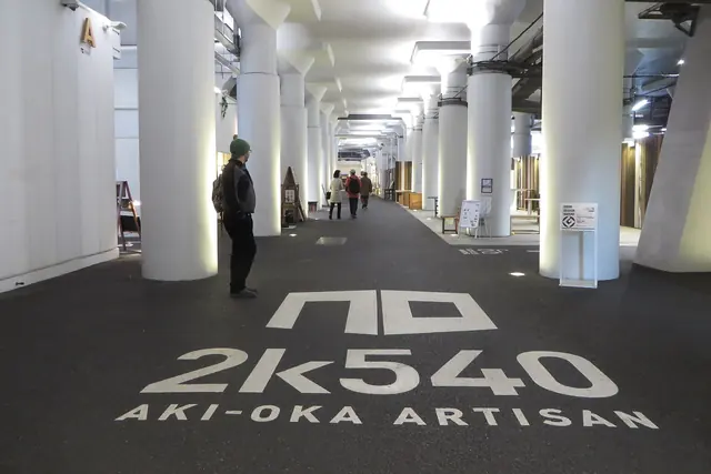A wide indoor walkway lined with thick white columns, with 2k540 AKI-OKA ARTISAN painted in large white letters on the dark floor