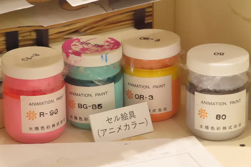 Four plastic jars of animation paint in pink, turquoise, orange and grey on a shelf, with a small handwritten card in Japanese in front of them
