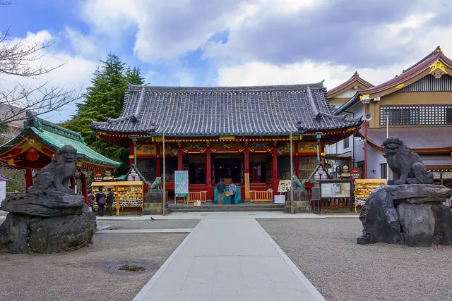 A shrine hall at the end of a stone-paved approach, with a stone guardian dog on a plinth at each side, under a blue sky with white clouds