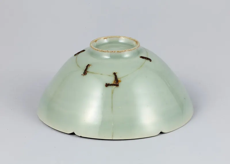 A pale green celadon bowl seen from below, with brown metal staples holding a crack