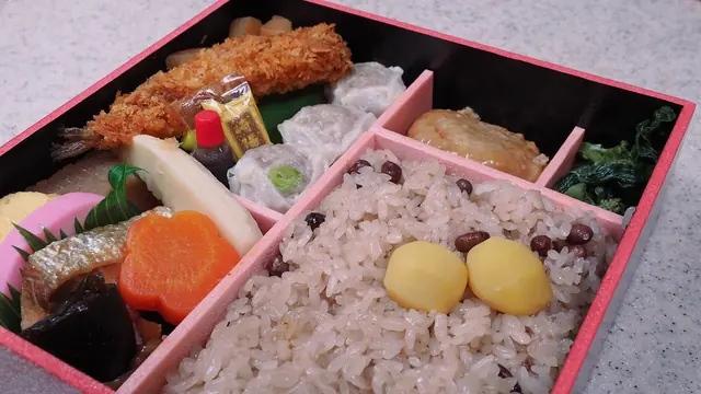 A bento box with red-bean rice and chestnuts, a fried prawn, dumplings, grilled fish and vegetables