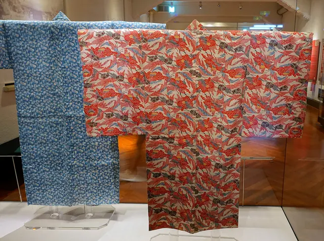 Two robes in Okinawan stencil dyeing, a red one with a bold flower pattern in front of a blue one, hung with their sleeves spread in a museum