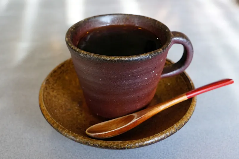 A dark reddish-brown unglazed cup of coffee on a matching saucer with a wooden spoon