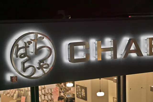 A backlit metal sign with a round logo of brushed Japanese characters beside large metal letters, above a lit shop window at night