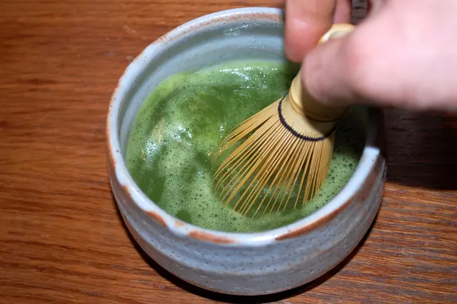 A hand moving a bamboo whisk through bright green frothy matcha in a pale grey bowl