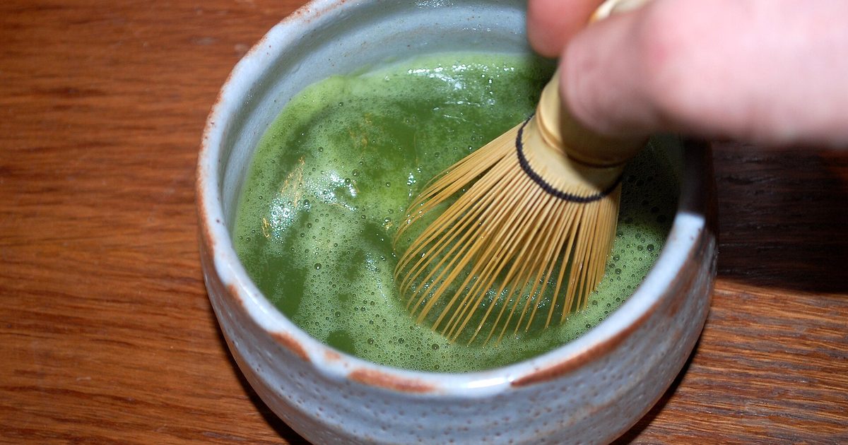 A hand moving a bamboo whisk through bright green frothy matcha in a pale grey bowl