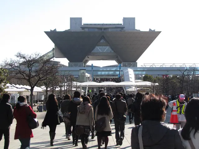 A crowd seen from behind walking past bare trees towards the inverted-pyramid Conference Tower of Tokyo Big Sight