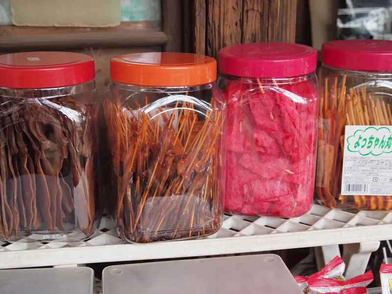 Four clear plastic jars with red, orange and pink lids on a shelf, filled with strips and skewered snacks