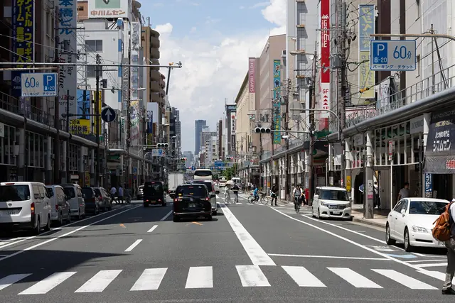 A wide street seen from a pedestrian crossing, lined on both sides with arcaded shops and signs, with cars and a parking sign