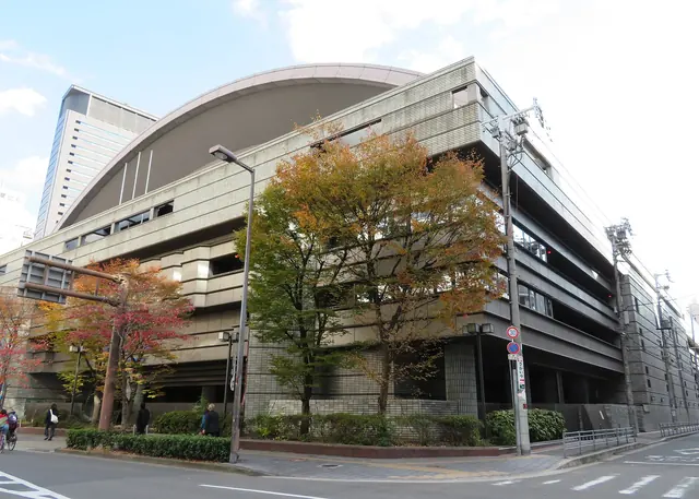 A large grey concrete arena with a curved roof on a street corner, with autumn trees in front