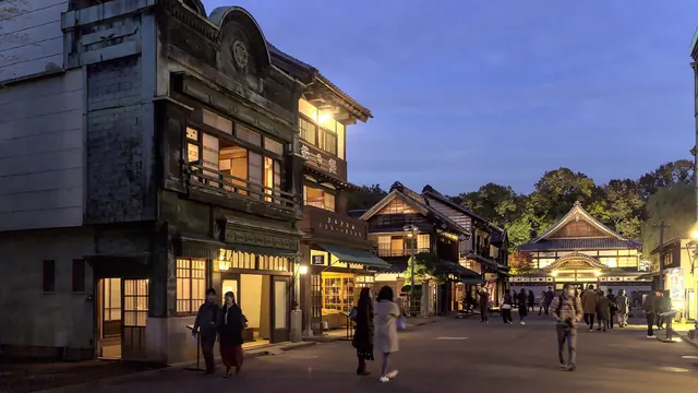 A street of old wooden shop buildings with their lights on at dusk and a few people walking