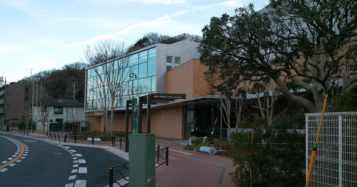 The entrance canopy of the Fujiko F. Fujio Museum, a glass and brick building beside a curving road, with bare trees in front