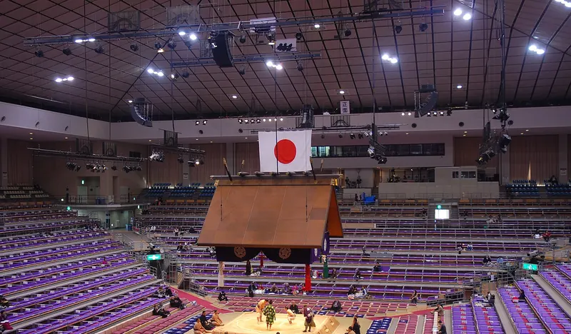 A sumo ring under a hanging wooden roof, ringed by floor cushions and rows of purple seats, early in the day with most seats still empty