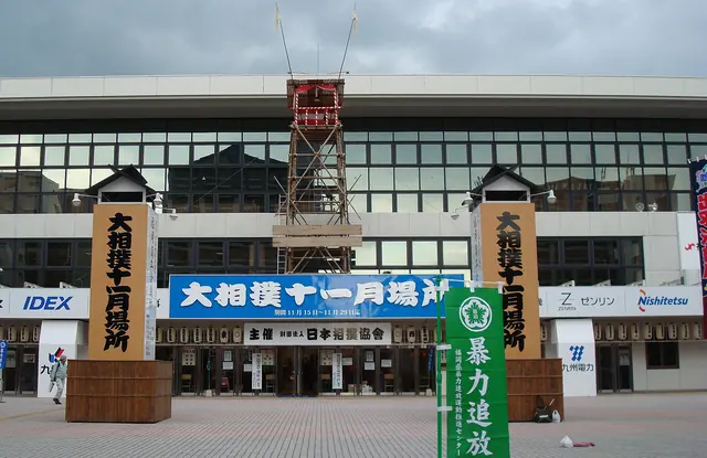 A tall wooden drum tower above the entrance of a glass-fronted arena, with two wooden pillars painted with black characters and a blue tournament sign