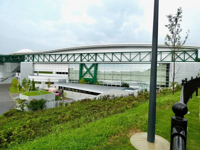 The outside of the building that holds Ghibli's Grand Warehouse, with a green steel frame along the roof, seen from a grassy bank