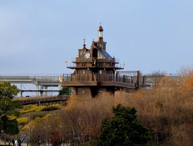 A brown tower with a clock and a small lantern top standing on a hillside above trees, with an elevated road behind