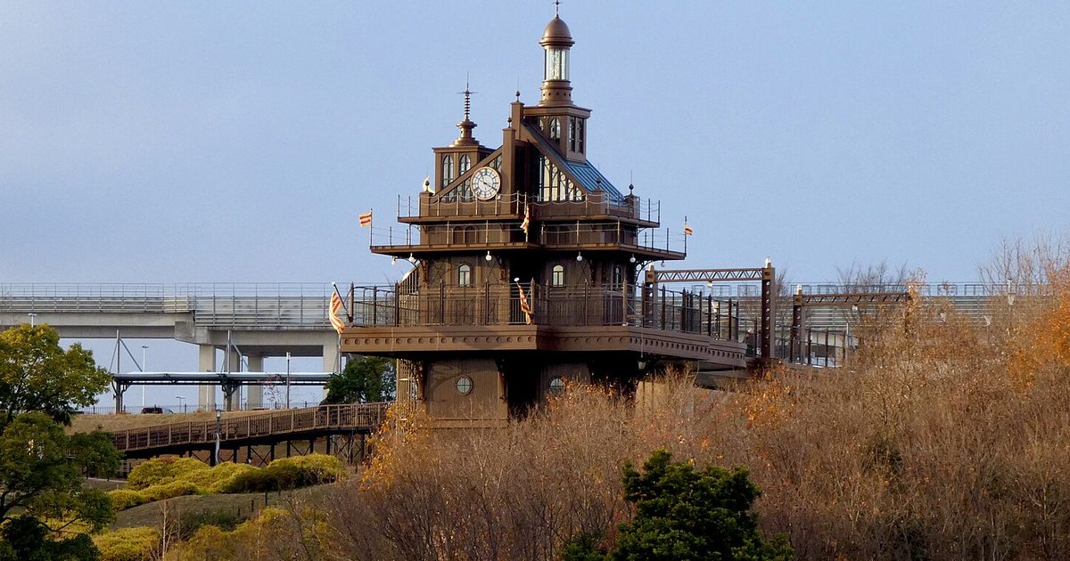 A brown tower with a clock and a small lantern top standing on a hillside above trees, with an elevated road behind