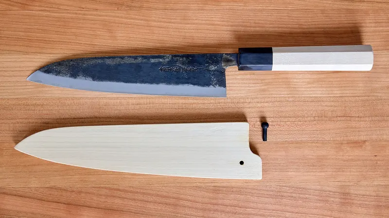 A Japanese chef's knife with a dark blade above its pale wooden sheath and pin, on a wooden board