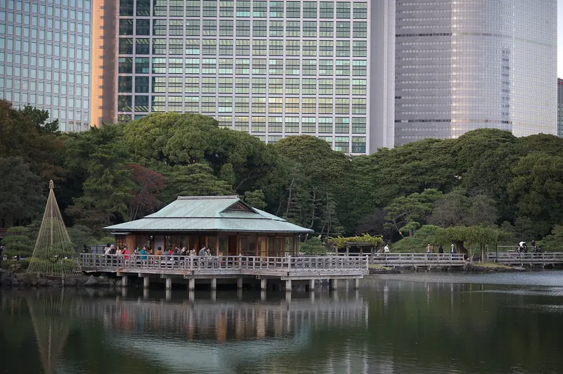 A teahouse on stilts over a pond with a long wooden bridge, trees and office towers behind