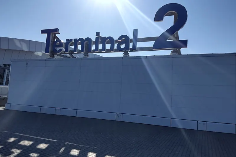 Large dark letters spelling Terminal 2 on top of a grey wall, with the sun shining from behind them in a blue sky