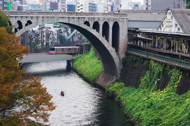 A concrete arch bridge over a river, with autumn trees on the left bank, a red-striped train crossing a low bridge beyond the arch and a station platform on the right