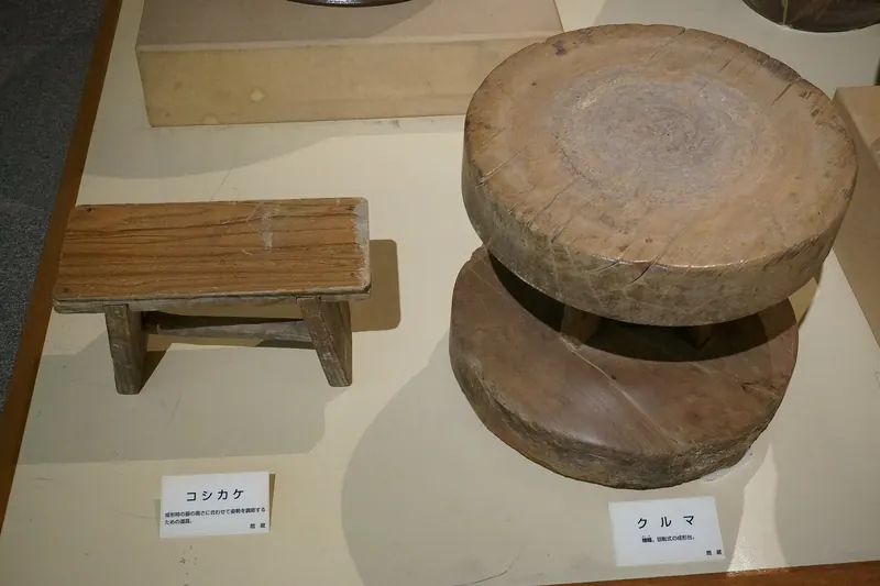 A heavy round wooden wheel and a low wooden stool on display in a museum case
