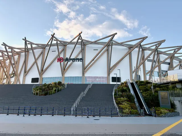 A white arena with a crown of slanting timber-coloured beams, wide stairs and planted slopes, under an evening sky