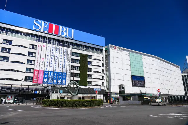 A white department store with a large blue SEIBU sign and a white PARCO building beside it, behind a roundabout with a hedge and a bronze sculpture, under a deep blue sky