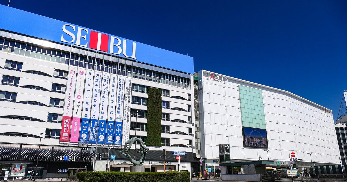 A white department store with a large blue SEIBU sign and a white PARCO building beside it, behind a roundabout with a hedge and a bronze sculpture, under a deep blue sky