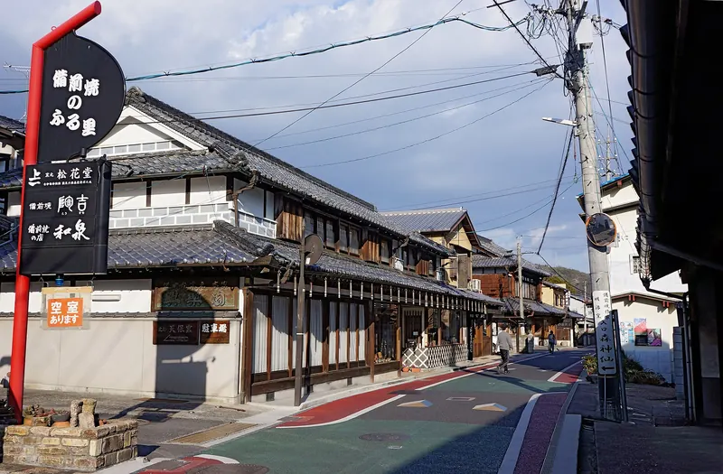 A street of old two-storey shops with tiled roofs and a red signpost on the left, under a blue sky