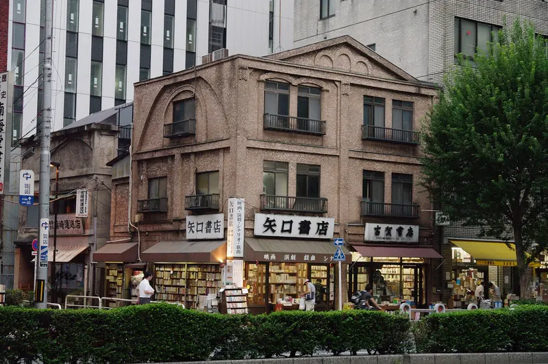A brown brick building from 1918 with arched upper windows and shelves of books along the pavement under its awnings, with modern office blocks behind
