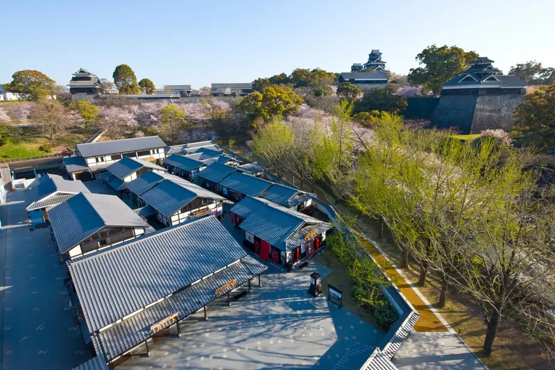 Rows of grey-roofed shop buildings among spring trees with the keeps of Kumamoto Castle behind