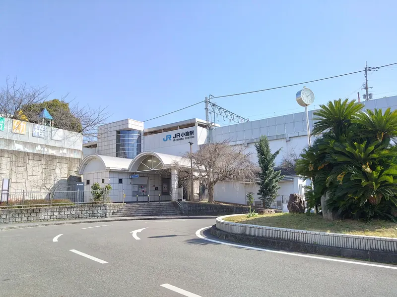 A white station building with arched roofs and a JR sign, behind a curved road and a planted roundabout