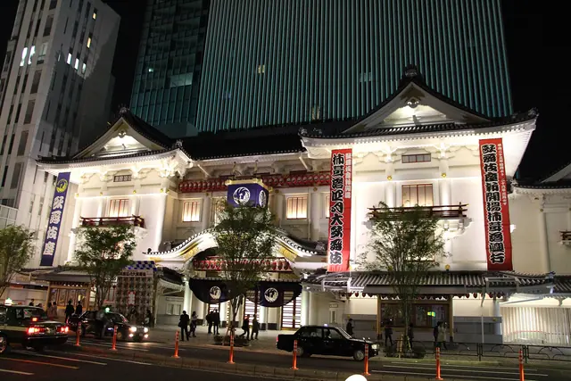 The Kabuki-za theatre lit up at night, with red and white banners on its white front and a dark glass tower rising behind it