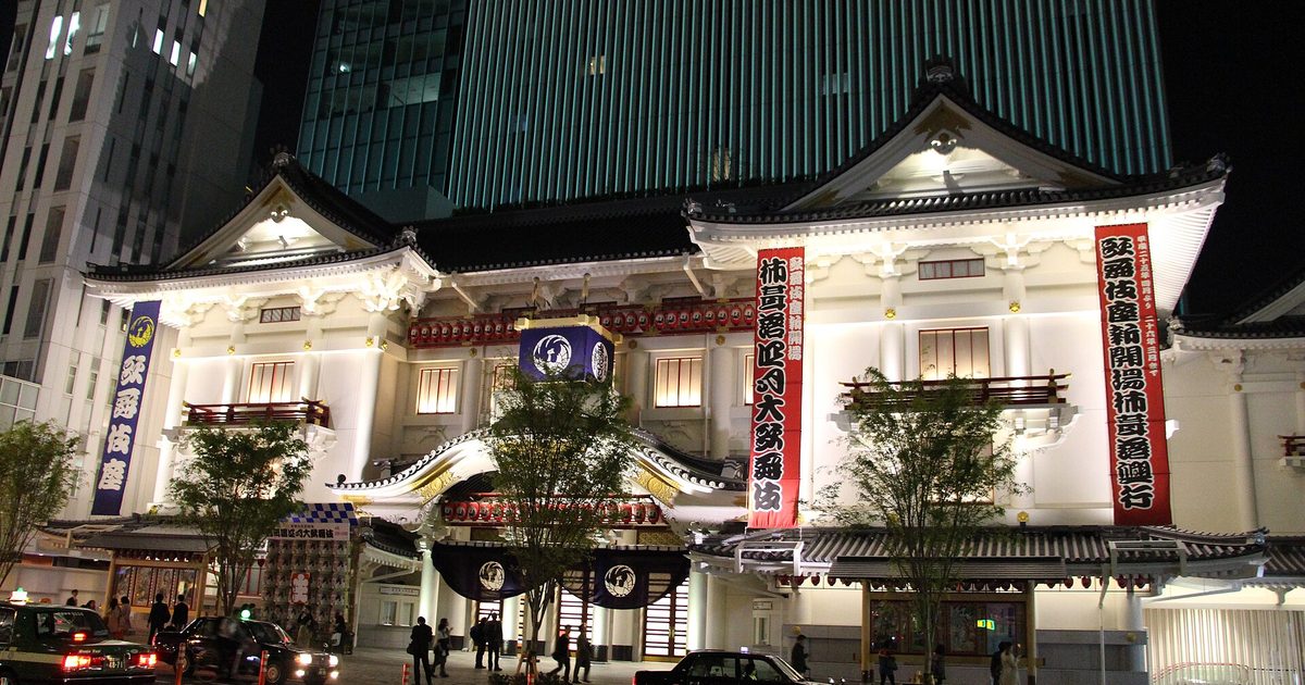 The Kabuki-za theatre lit up at night, with red and white banners on its white front and a dark glass tower rising behind it