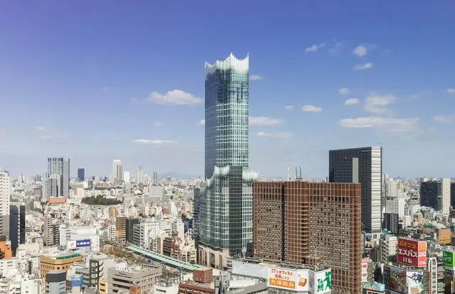 A tall glass tower with a wave-shaped top rising above the Shinjuku skyline under a blue sky