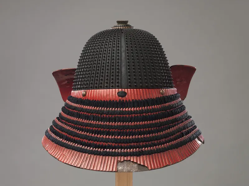 A riveted black iron samurai helmet with a red-laced neck guard and red side flaps on a wooden stand
