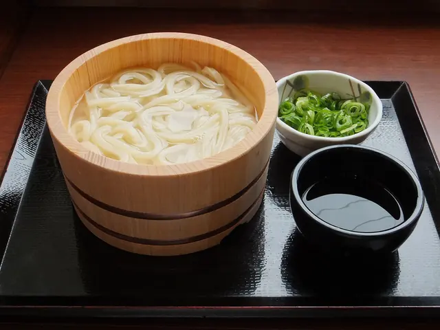Udon noodles in a wooden tub with a cup of dipping sauce and a small bowl of spring onions on a tray