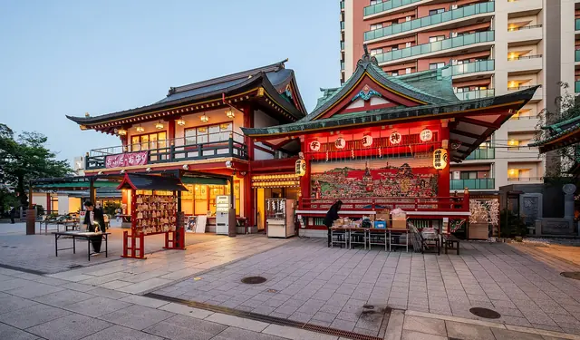 Two vermilion shrine buildings lit at dusk on a stone-paved courtyard, with an apartment block behind
