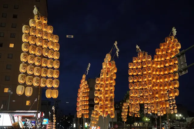 Tall bamboo poles hung with rows of lit paper lanterns against a night sky, with city buildings behind