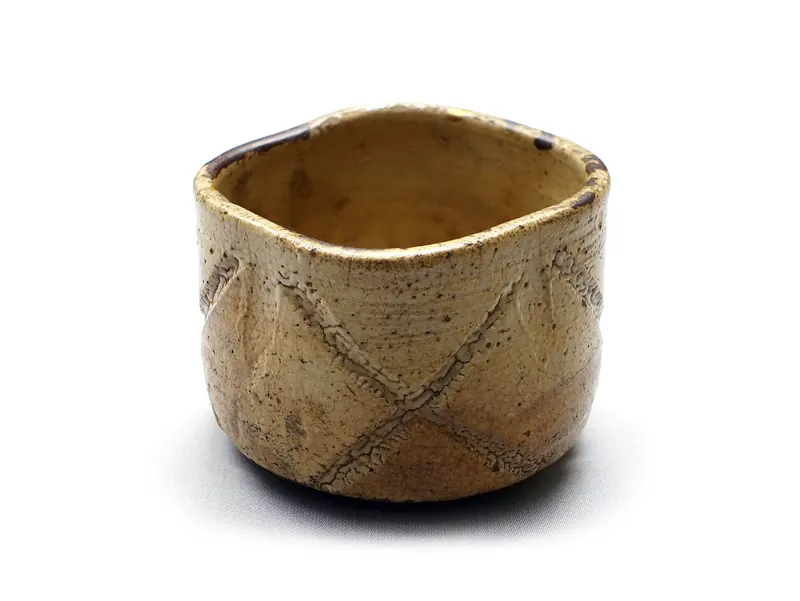 A beige tea bowl with a slightly uneven rim and crossed lines pressed into its side