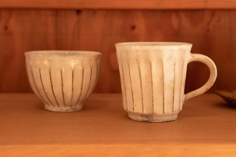 A small cup and a mug in white glaze with carved vertical lines, on a wooden shelf