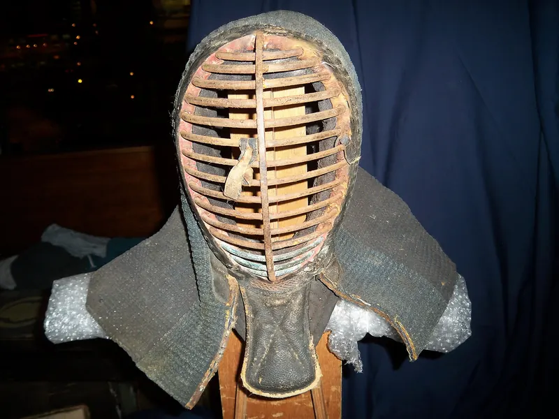 An old kendo head guard with a metal grille and padded cloth flaps on a stand against dark cloth