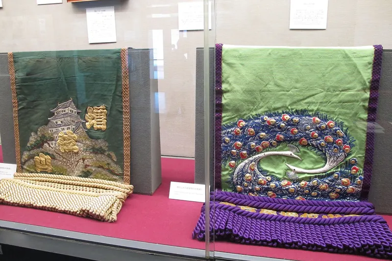 Two embroidered ceremonial aprons in a glass case, one dark green with a castle, the other pale green with a large coiled pattern in jewel colours, each with a thick fringe along the bottom