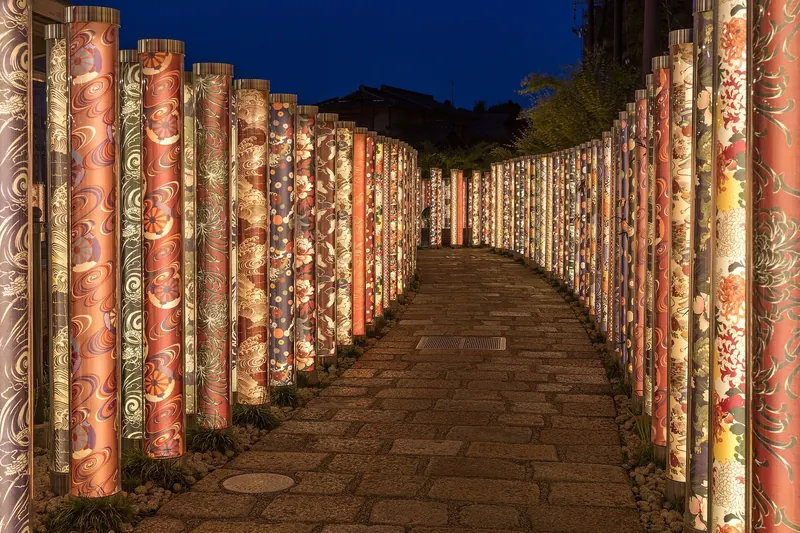 A stone path curving between tall illuminated poles wrapped in kimono-fabric patterns at dusk
