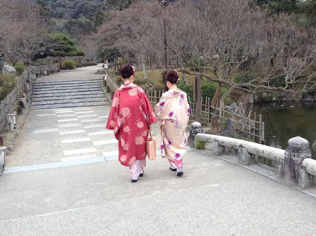 Two people in red and pink kimono, seen from behind, walking along a paved path towards stone steps in a garden