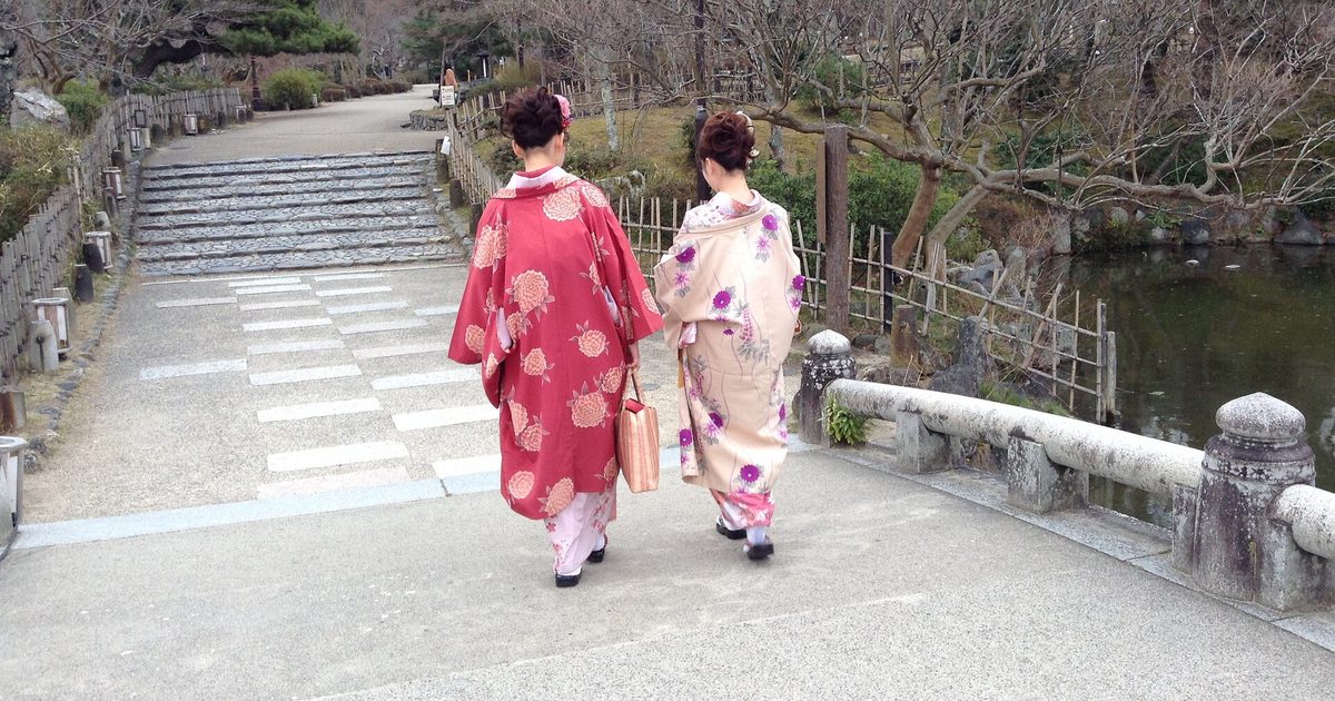 Two people in red and pink kimono, seen from behind, walking along a paved path towards stone steps in a garden