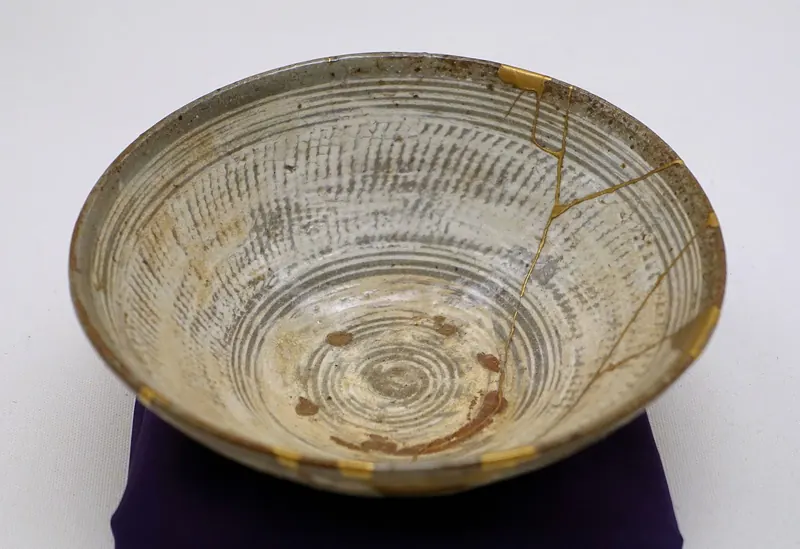 A shallow grey stoneware bowl with incised rings and thin gold lines along its cracks, on a purple cushion