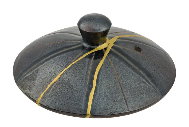 A dark grey teapot lid with a knob on top, its cracks joined by bright gold lines