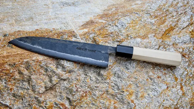 A hand-forged Japanese chef's knife with a dark, rough-finished blade, a polished edge and a pale wooden handle, lying on a rock